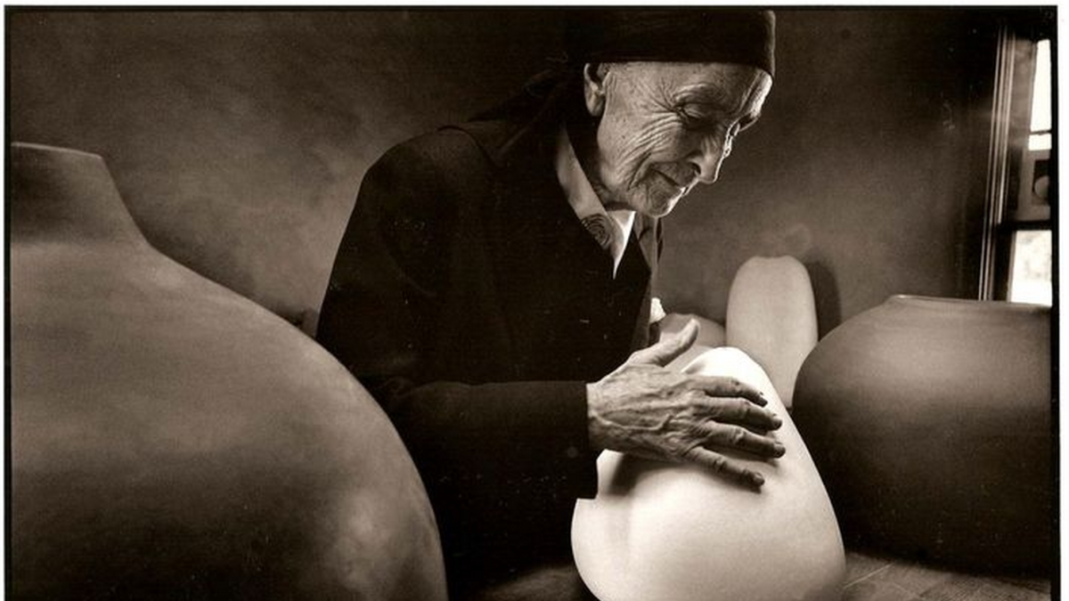 Black and white photo of the artist Georgia O'Keeffe late in life, dressed in black loose clothing with a black head wrap with her hands placed upon a white curvilinear abstract sculpture about the size of a large pumpkin; she is in profile looking down at her work. 