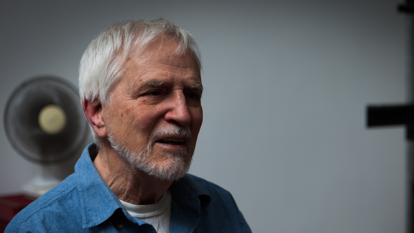 Close up photo portrait of Lennart Anderson in his studio, with gray beard, gray hair, blue button up shirt with white t shirt in the middle of speaking.