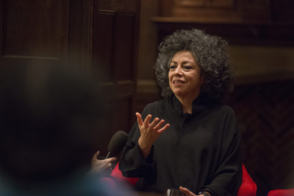 Color photograph of the artist Doris Salcedo in a dark classroom, dressed in dark clothing, holding her hand out  in a gesture as if inviting conversation; Salcedo has dark, curly hair with some silvering, and she looks poised as if making a point or a statement to her audience. 