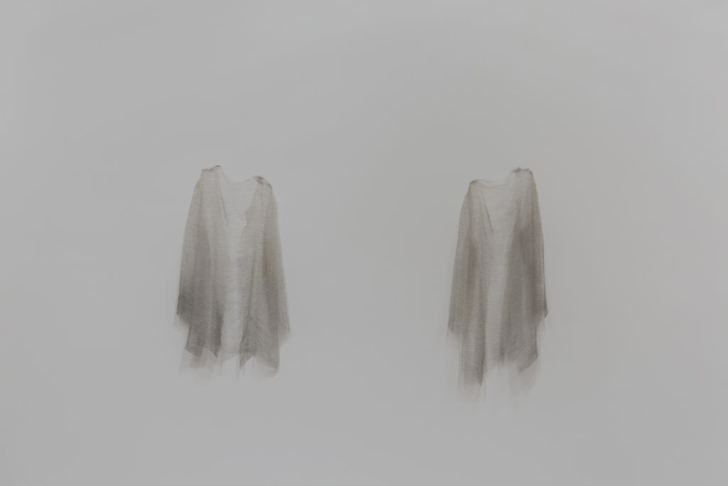 A photograph of two ethereal looking cloaks hanging from a wall. The cloaks are woven from silk thread and steel straight pins.