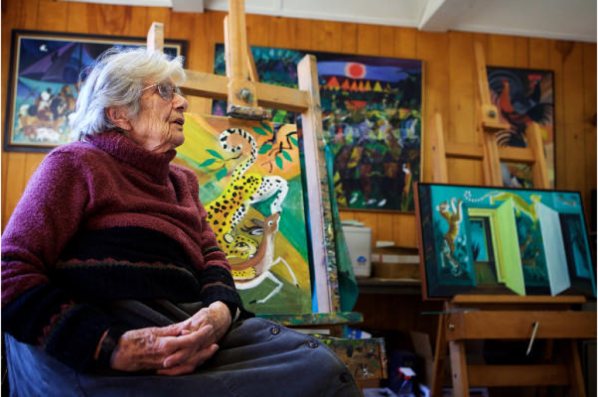 Chromatically colorful and bright photograph of an elder woman with silver hair sitting justified left in the frame, looking to the right, in an oversized dark purple sweater with her hands on her lap and multiple paintings in the background. 