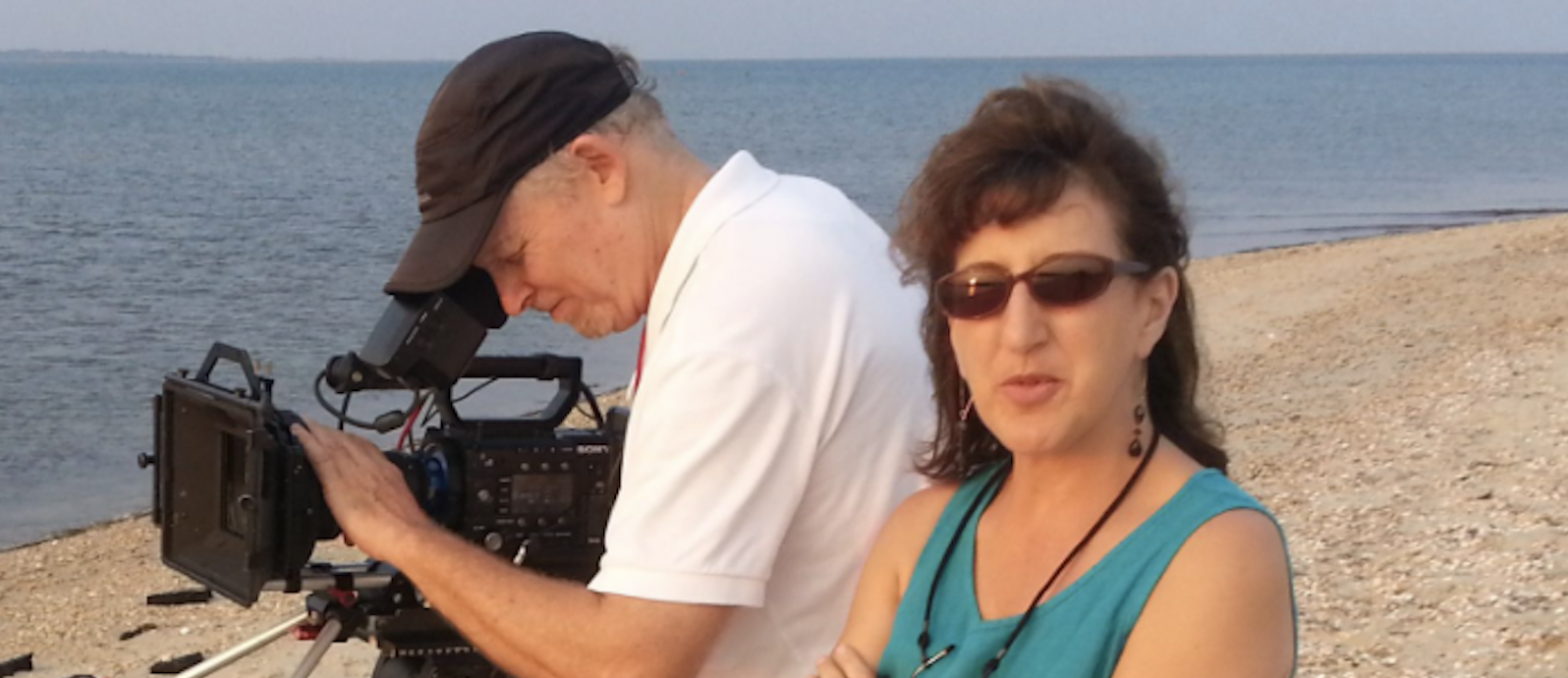 color photograph of filmmaker Andrea Torrice standing on a beach in blue sleeveless top and sunglasses with arms crossed looking at the viewer with cameraman in white t shirt behind looking into large black camera on tripod with ocean to the left