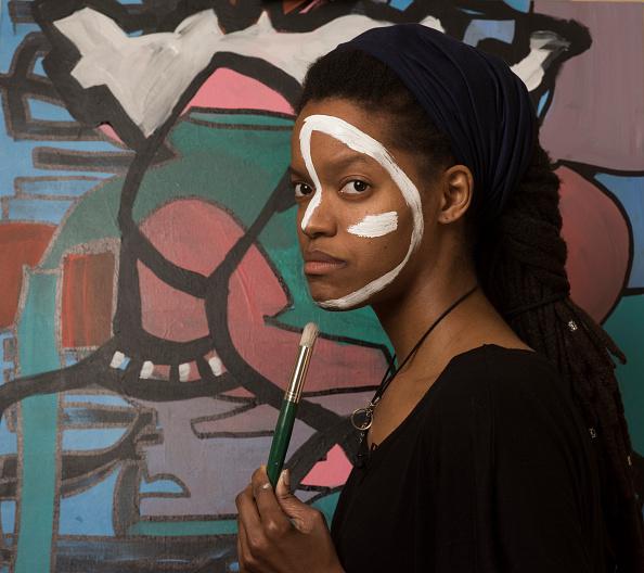 Full color photograph of the young artist Erika Marie York; the artist is posed in profile from the waist up, facing to the left with with eyes turned to the viewer; she is holding a brush vertically, and her face is painted with a white linear shape that wraps around her forehead and onto the ridge of her nose evoking primitive face painting and mimicking one of her abstract paintings that is set behind her. 