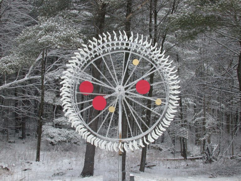 Round aluminum mobile with spokes like a wagon wheel that has three red circles attached to it, seen from a distance mounted to a tree in a field of white snow, winter.