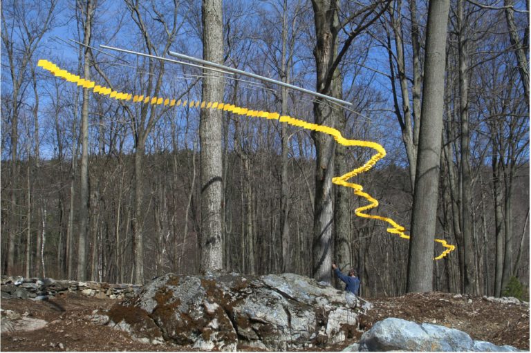 Long, yellow kinetic serpentine sculpture winding between tall, skinny trees like a snake, with blue sky and snow on the ground.