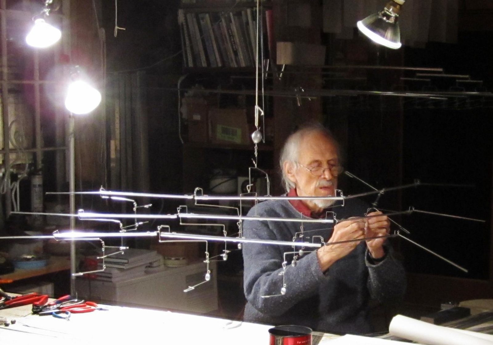 Older man with white whispy hair sitting at a table with bright lights to the right, working on an aluminum mobile hanging from the ceiling