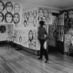 Black and white photo of a woman in gray walking away from the viewer in an interior of a room with the walls covered with portrait drawings