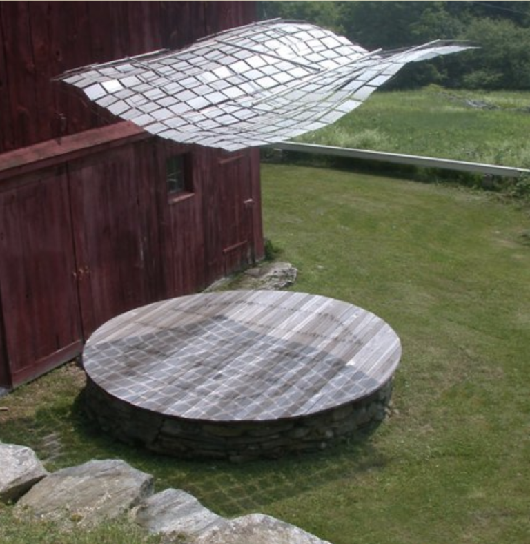 View from above looking down on a shiny metal fabric of small squares waving in the wind, suspended over a large wooden platform in a field of grass in front of an old red barn