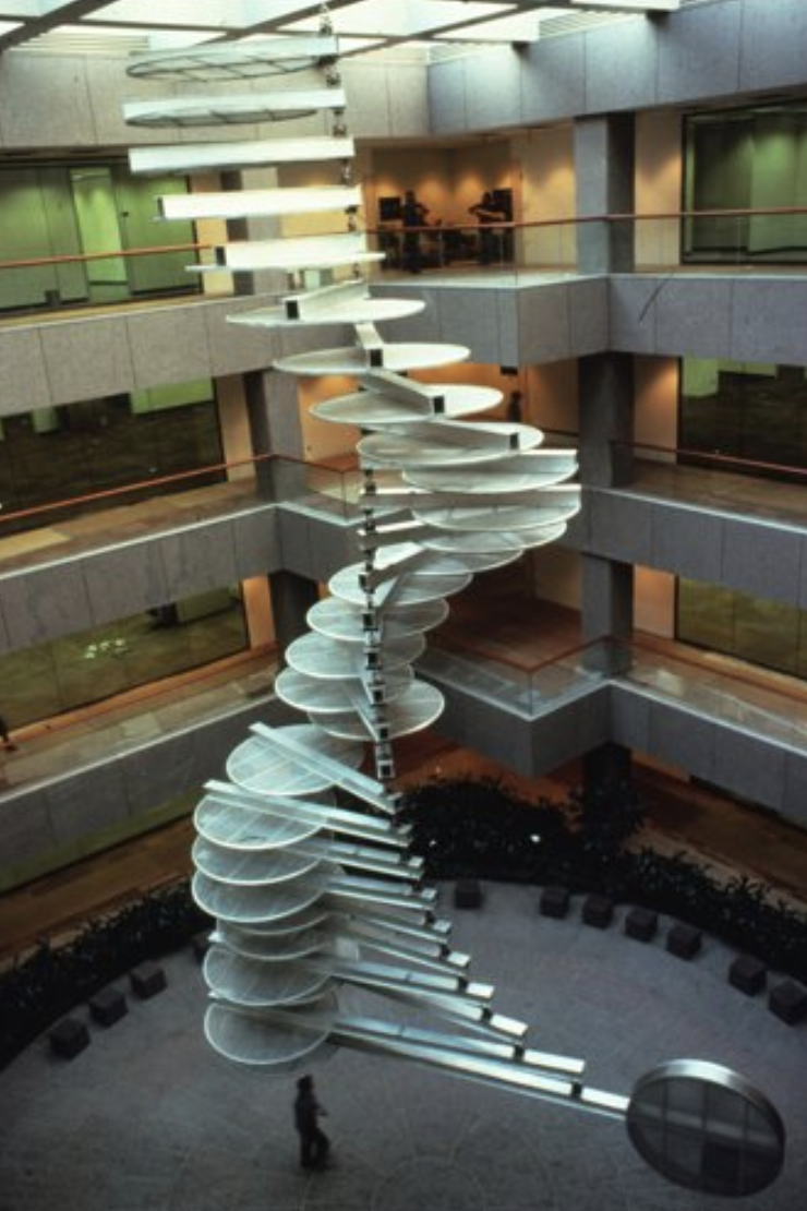 View of an interior public lobby of a tall, sinuous metal construction in the shape of spiraling vertebrae constructed of horizontal discs and metal armatures connecting to a floor to ceiling metal tie rod.