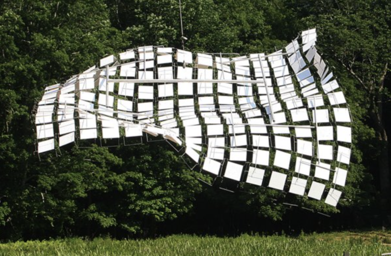 Shiny fabric made of small squares of aluminum waving in the wind suspended horizontally over a field of green grass with a dense, dark forest in the background