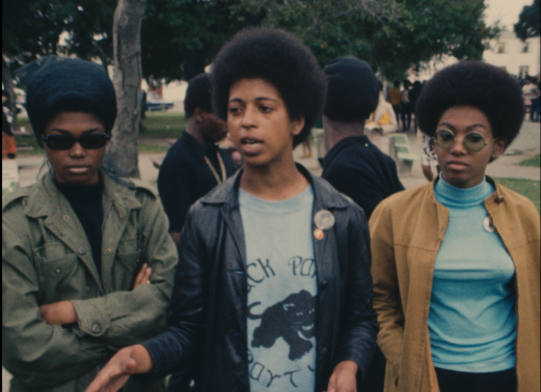 Three women stand next to one another at a 1968 Black Panther demonstration in Oakland, California in a still from Agnès Varda's 1968 film "Black Panthers." The woman in the center is speaking and gesturing with her hands; she has an afro and is wearing a leather jacket open over a Black Panther tee-shirt. The woman to her left has on a head covering and dark glasses; her arms are crossed in front of her. The woman to her right has an afro and is wearing a blue turtleneck and lightly-tinted glasses; her hands are tucked into the pockets of her tan jacket. Two men are standing behind them and looking back at what looks like a crowd of people in the distance.