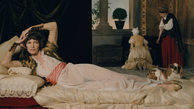 Color still from Agnès Varda's 1988 film “Jane B. par Agnès V.” showing Jane Birkin in period costume in a tableau vivant of Titian's 1538 painting, Venus of Urbino. The main female figure is in the foreground, reclining horizontally across the picture frame. She's on her side, facing the viewer with a direct gaze, propped up against a large pale yellow silk pillow on the left with her hands poised behind her head. She has long brown hair, her elbows are flared out. She is wearing a long, loose, off-white translucent dress with a pale red sachet around the waist. Two women in long period dresses are beyond, focusing on a task, and a dog with mottled white and brown fur and long, soft ears sits at her feet. Its gaze is also trained directly at the viewer.