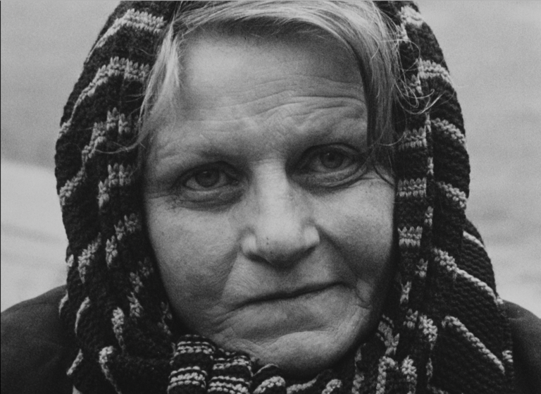 Black and white portrait photograph of an older woman with her full face filing the frame, her soft features and eyes are turned slightly up and she is looking away beyond the viewer; she has a loosely knit dark patterned scarf wrapped around her head so that only the flesh of her face is visible, with small locks of hair framing her forehead; her eyes are in the shadows of her heavy brow and she is lost in thought.