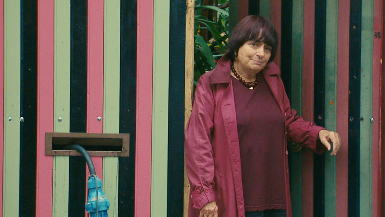 Late life color photograph of Agnes Varda leaning against two doors that are patterned with vertical stripes in alternating hues of muted green, red, and black; Agnes has short cropped dark hair with red accents, her head is tipped slightly to the right, and she is wearing a loosely fitted maroon jacket, a necklace of dark red beads, and a loose, dark red knitted sweater. She is leaning agains one of the doors with her hand prominently placed toward the right of the frame; a bronze mail slot is on the left with a blue electrical cord protruding from it.