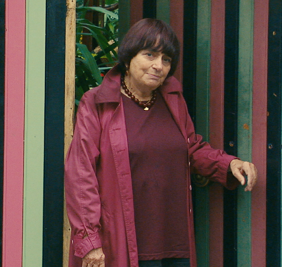 Late life color photograph of Agnes Varda leaning against two doors that are patterned with vertical stripes in alternating hues of muted green, red, and black; Agnes has short cropped dark hair with red accents, her head is tipped slightly to the right, and she is wearing a loosely fitted maroon jacket, a necklace of dark red beads, and a loose, dark red knitted sweater. She is leaning agains one of the doors with her hand prominently placed toward the right of the frame; a bronze mail slot is on the left with a blue electrical cord protruding from it.