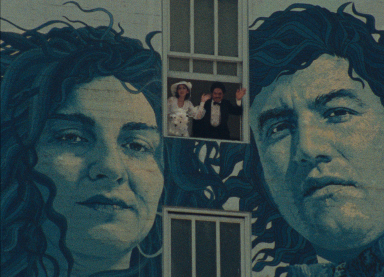 A man and a woman dressed for a wedding lean out of a window. Their figures are overshadowed by a huge mural of a man and a woman on the wall of the building the couple are in. The portraits in the mural and detailed and highly realistic.