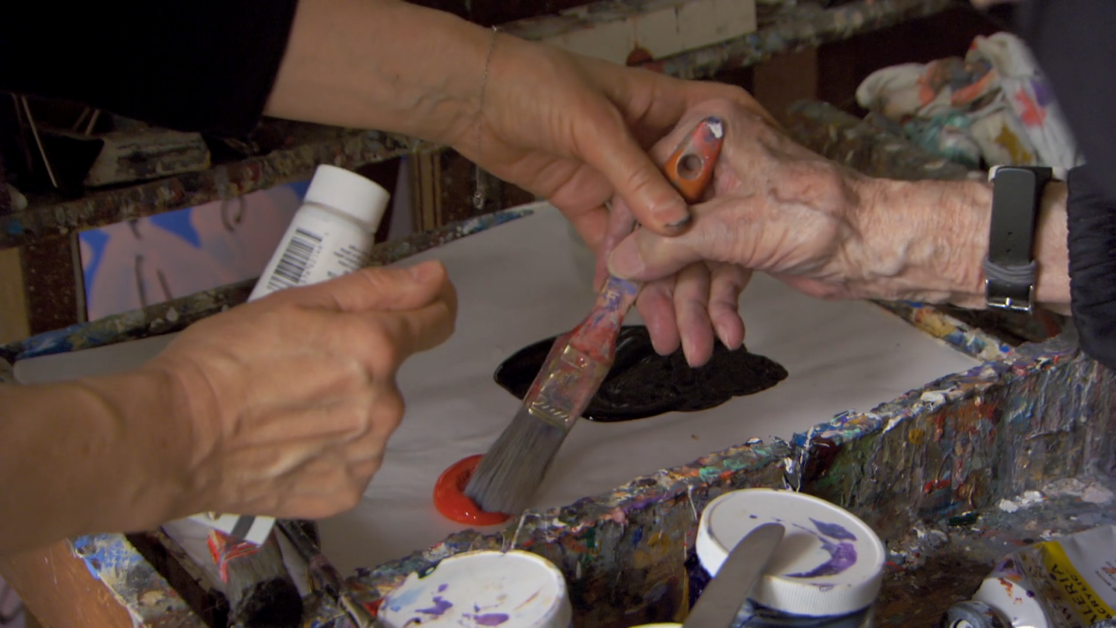 In a still shot from "Serge Hollerbach: A Russian Painter in New York," Olga Nenazhivina's hand guides Serge Hollerbach's hand, in which he is holding a paintbrush, to the right pile of red paint on his palette.
