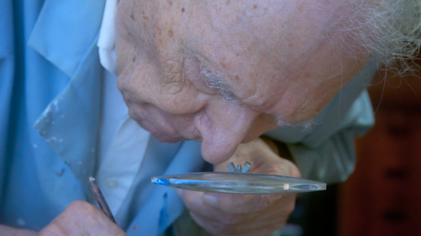 In a still from "Serge Hollerbach: A Russian Painter in New York," Serge Hollerbach's head is seen in extreme closeup as he looks down to work on touching up a painting. He's looking through a thick magnifier, which he holds in his left hand. In his right hand, which is only partially visible, he holds a paintbrush.