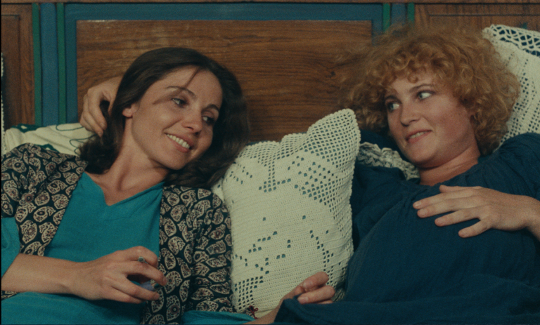 Thérèse Liotard, who has straight dark hair, and Valérie Mairesse, whose red hair is curly, lounge next to one another on a bed in a still from Agnès Varda's 1977 film, "One Sings, the Other Doesn't." They are propped up against pillows with lace covers. Both women are relaxed and happy. Thérèse Liotard, who plays Pauline, is smoking. Valérie Mairesse, who plays Pauline, is visibly pregnant. Her hand rests on her belly.