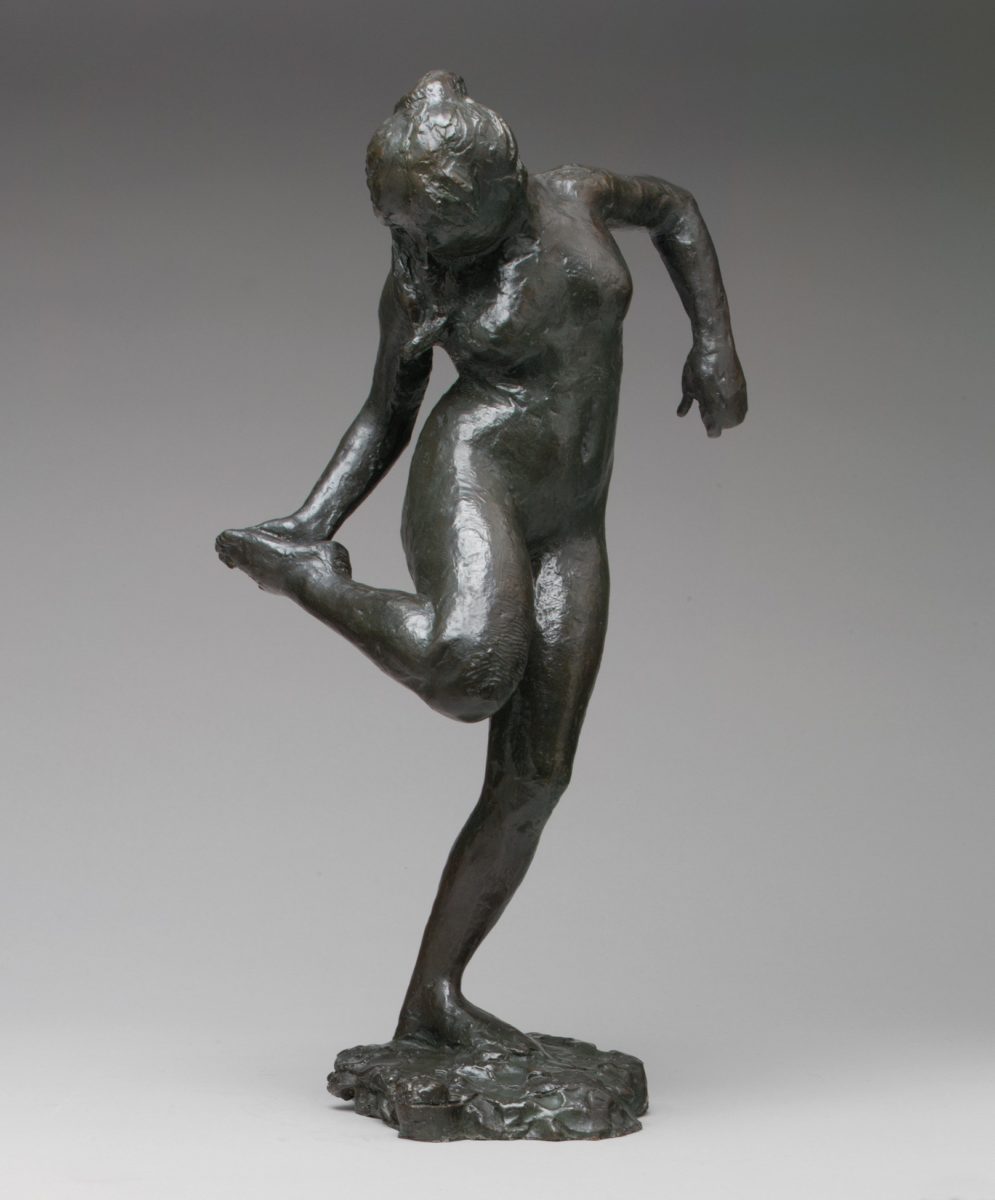 Dark bronze full figure sculpture of a woman standing on one leg with head turned away and looking down at the sole of her foot which she is holding upturned in her right hand. Her other arm with elbow turned up and away.