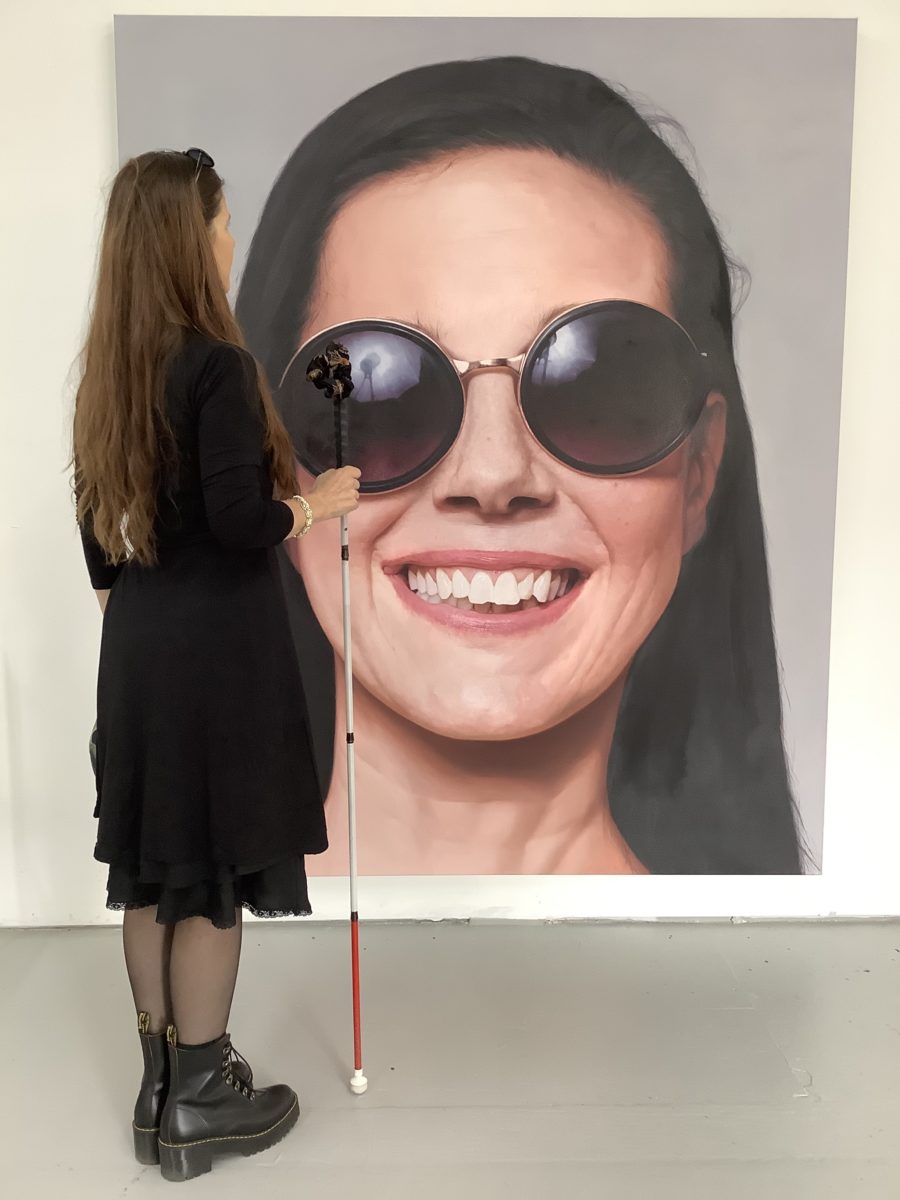 Image of a large scale photographic portrait of a woman looking at the viewer smiling with round dark sunglasses with a woman seen from behind in full figure with back turned, looking at the portrait; the woman standing with back turned is holding a walking cane with a red tip used by blind persons