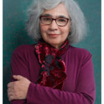 Full color three quarter portrait photograph of a woman with shoulder length silvering hair, arms crossed, in a muted purple long sleeve shirt and shimmery red and black scarf; the woman is wearing dark rimmed plastic glasses, is looking directly at the viewer with a slight, knowing smile.