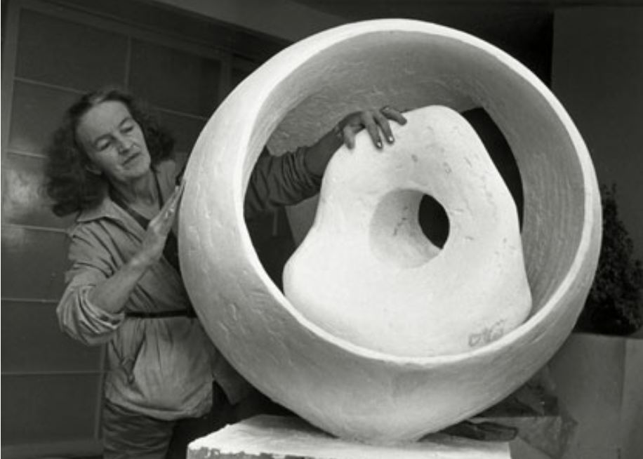 A black and white photo fo a woman standing cropped from the waist up next to a large, round white sculptural form. The large white form is hollowed out in the middle, inside of which is placed an amorphous organic form with a hole through the center of it. The woman's right hand is touching the round sculpture and her left hand is reaching into the sculpture draped over the interior amorphous form.