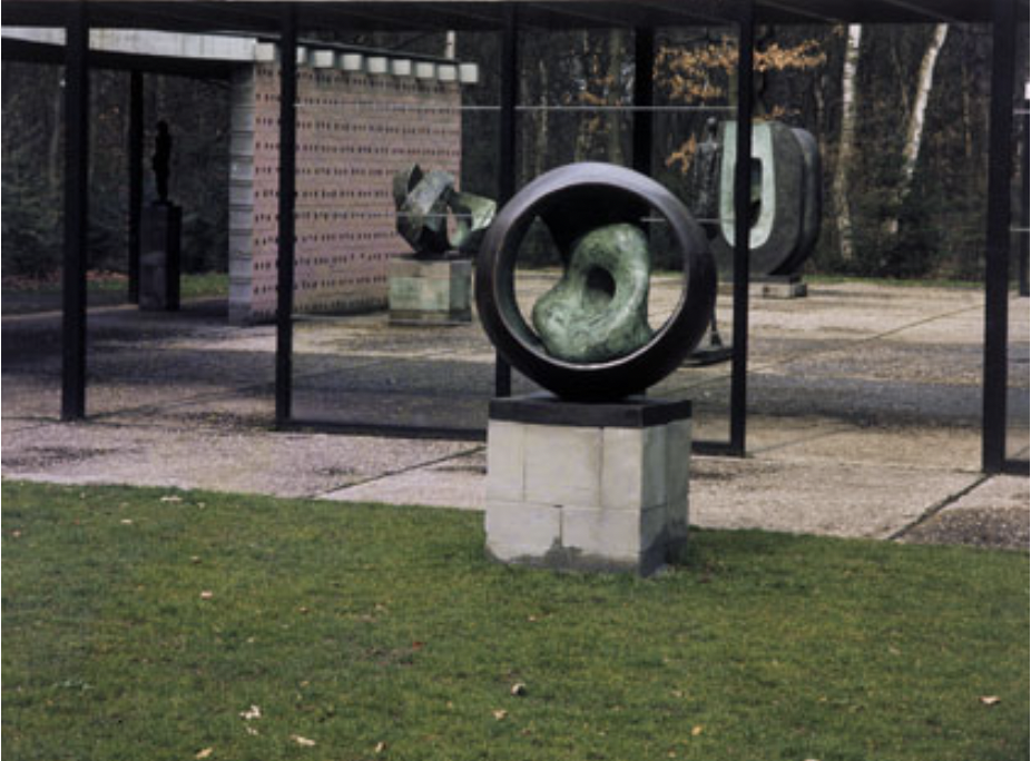 Color photograph of an outdoor courtyard with concrete pavers and closely cropped grass. At the center of the photo, sitting in the grass, is a square stone plinth with a round, abstract bronze sculpture placed on top of it. The bronze sculpture is in the shape of a ring, inside of which is placed an amorphous organic form that looks like green patina'd stone with a hole in the center of it. Several more abstract sculptures can be seen in the background.