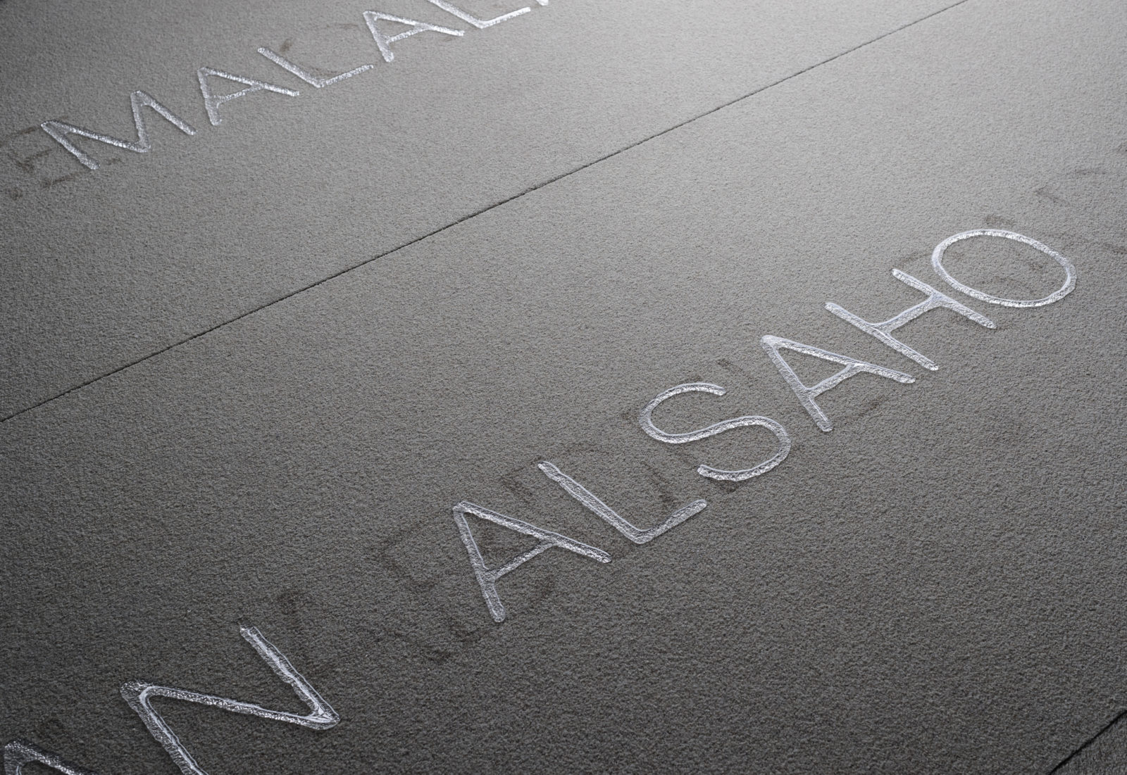 Black and white photograph of a soft, textured surface with layers of simple, san serif capitalized font moving diagonally from bottom right to top left. Dark font that is barely visible lays underneath lighter font that is more prominent and clearer, spelling the name “ALSAHO.”