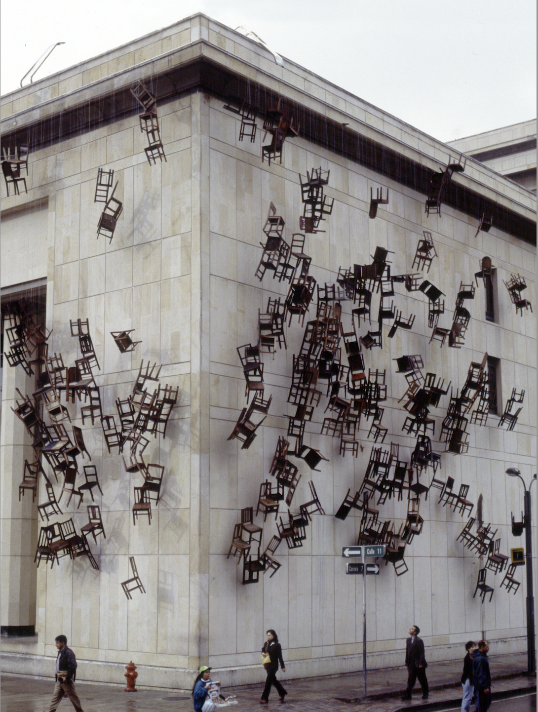 Color photograph of the exterior of a light grey municipal building, seen from slightly above. Thirty to forty dark wooden chairs hang are being lowered from the eaves of the building, making it look as though the building is being swarmed by large bugs or animals. A dark, wet sidewalk runs left to right across the bottom of the photo where half a dozen people are captured frozen, walking purposefully.