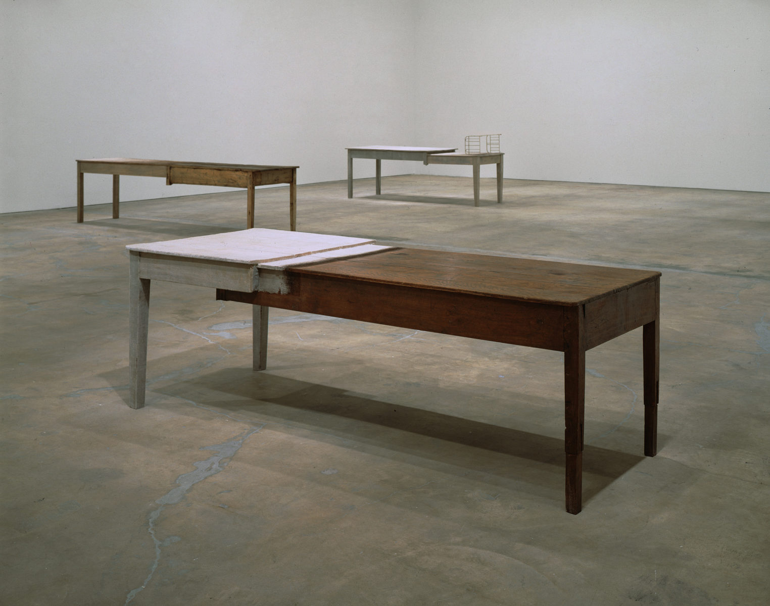 Color photograph of the interior of a vast, spare room with white walls and unfinished concrete floor. Three rectangular tables are placed parallel to each other evenly spread across the expanse of the room. Each table is composed of one half of a white table and one half of a natural wood table, carefully spliced in the middle with the white table overlapping the natural wood table.