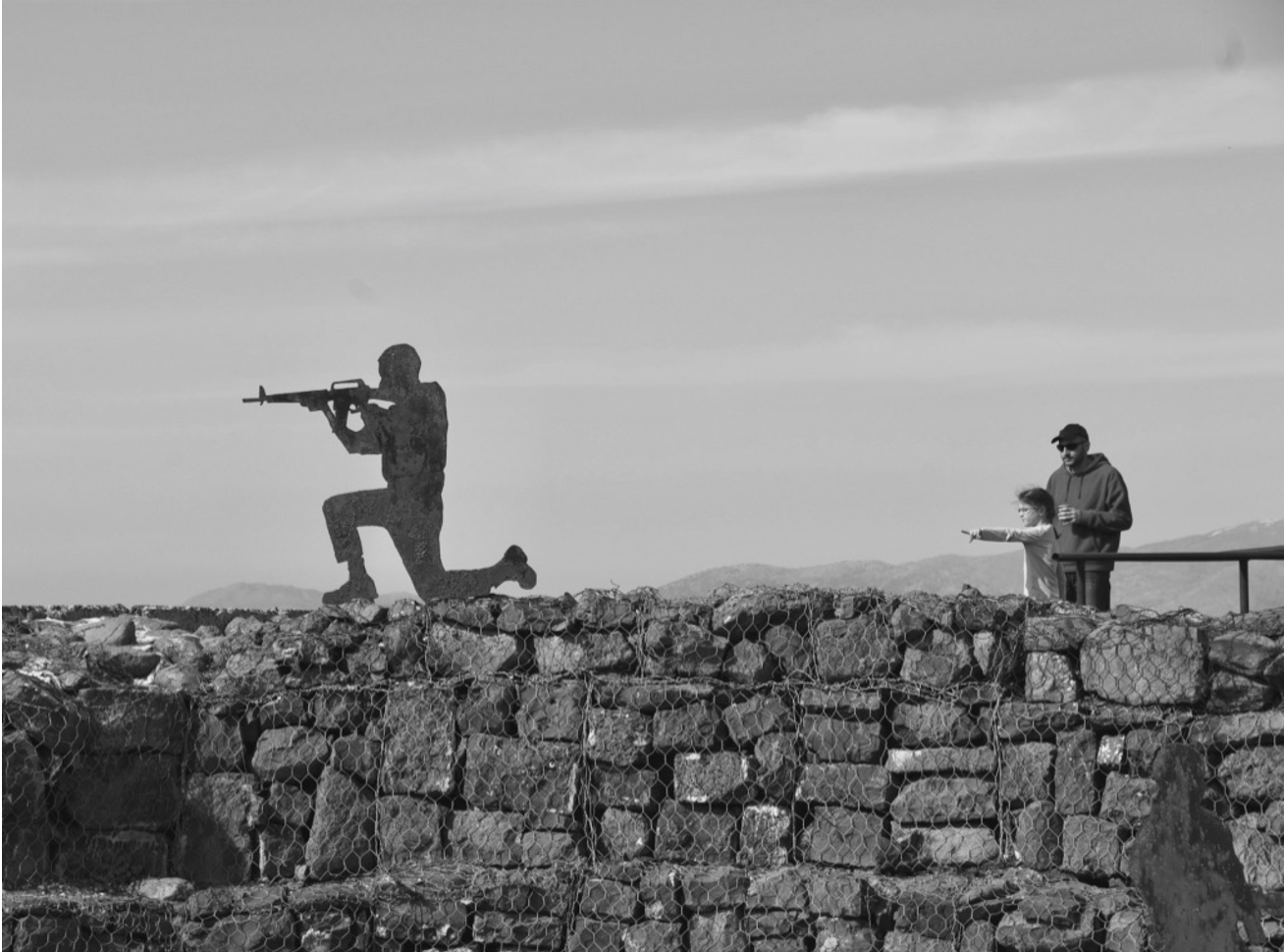 A black and white photograph. There is a wall made of stone with fencing in front of it. On top of the wall is a metal cutout of a man in silhouette holding a rifle and posed for stability with one knee bent at a 90 degree angle in front of him and one knee on the ground. A man and child stand at the wall. The child points toward the silhouetted figure. Half the photograph is of the still sky.