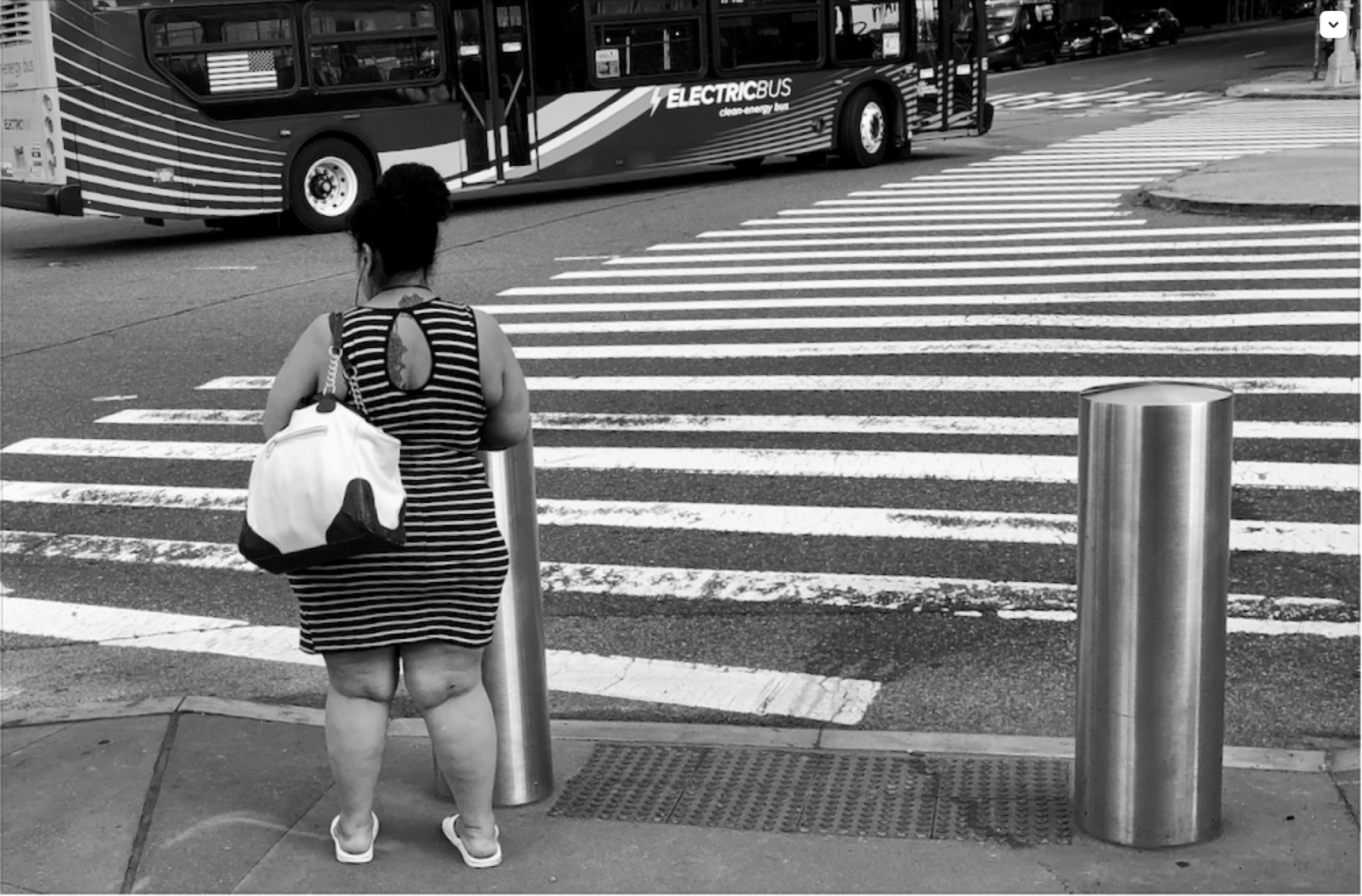 A black and white photograph of a woman seen from behind in a striped dress standing on a streetcorner by a sidewalk. A bus parked on the other side of the street also sports stripes.