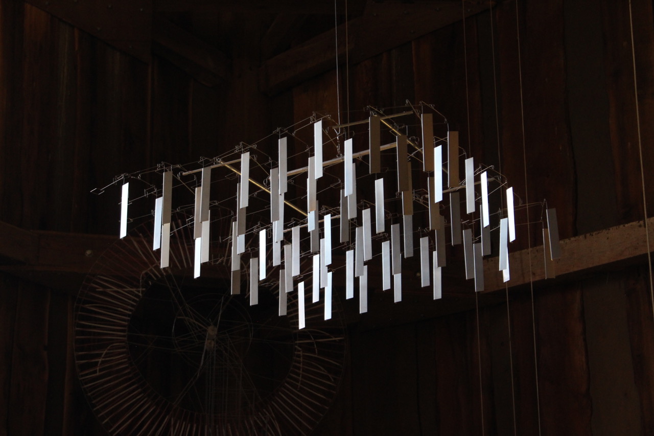 A chandelier-like network of silver rectangles hangs from a square armature of wire and is suspended from the ceiling in a dark, timbered space.