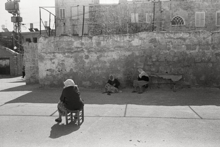 A woman in a white headscarf sits in a chair, her back to the viewer, in a slab of sunlight, facing a stone wall.