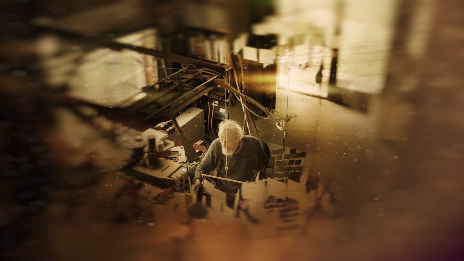 A sepia toned still shot from a film of an older man in his art studio, surrounded by his kinetic works of art. We are looking down into the studio from a slightly elevated point of view.