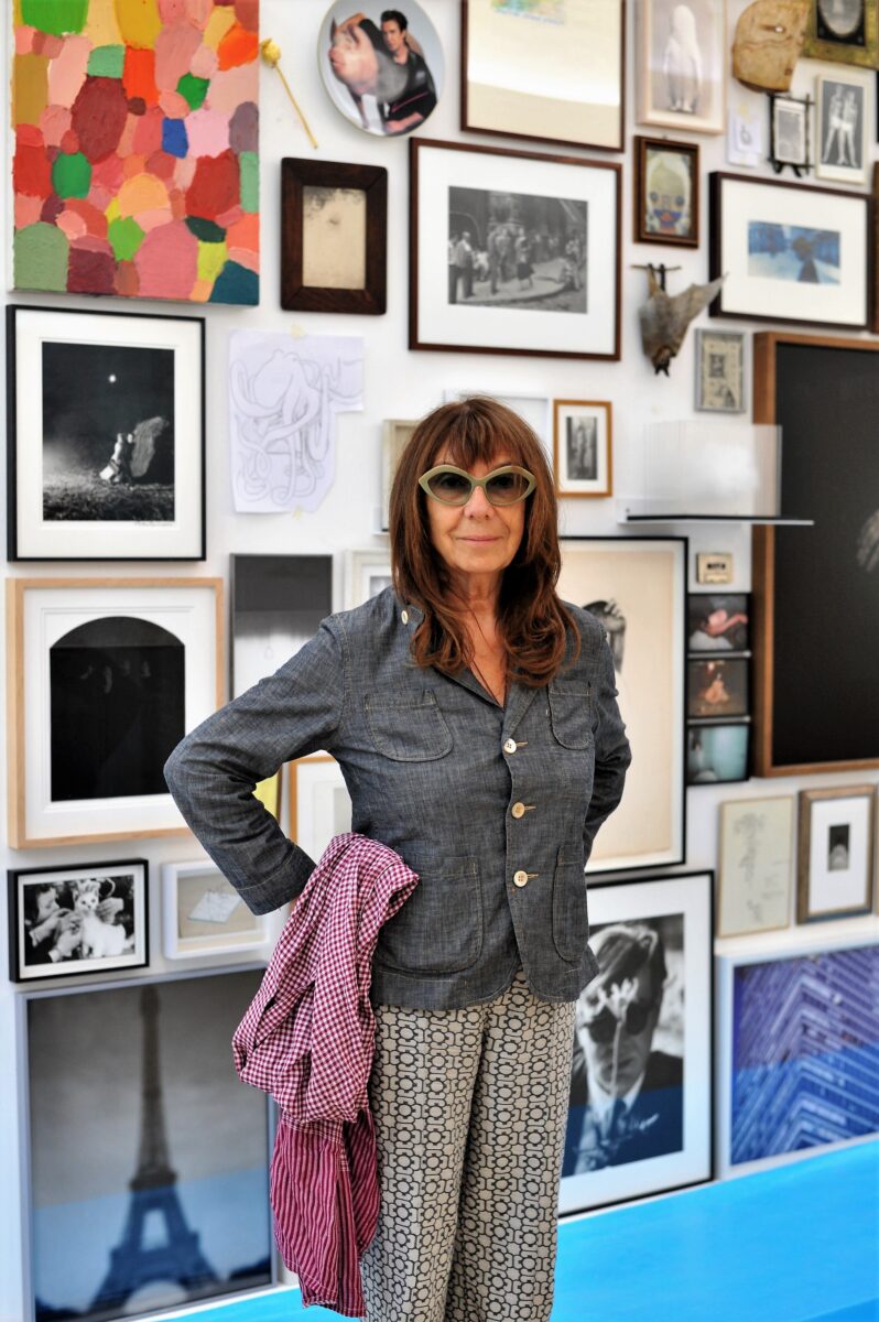 A photo of the artist Sophie Calle in front of a wall covered with photos, sketches, masks, and even a taxidermy bat. Calle is wearing sunglasses and patterned pants. Her hands rest on her hips, and her lips turn upward in a faint smile.
